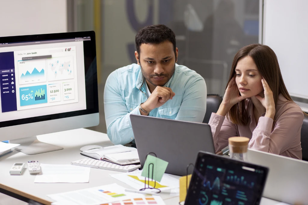 Two professionals collaborate on a business project in a modern office environment surrounded by multiple digital devices and data visualizations.