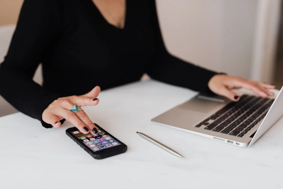 A person in professional black clothing works at a white desk with a laptop, smartphone, and notebook in an indoor office or workspace setting.