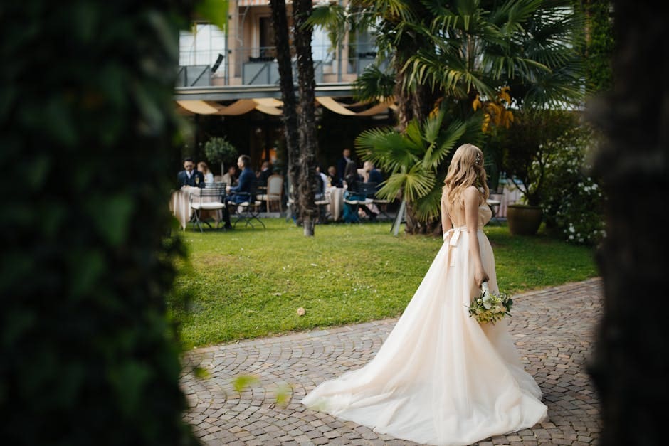 A bride in a flowing white wedding dress walks through a lush garden venue with guests visible in the background.