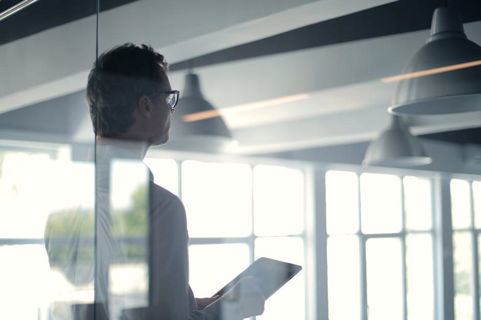 A professional wearing glasses holds a laptop while standing in a modern, naturally-lit office space with glass partitions and exposed ceiling beams.