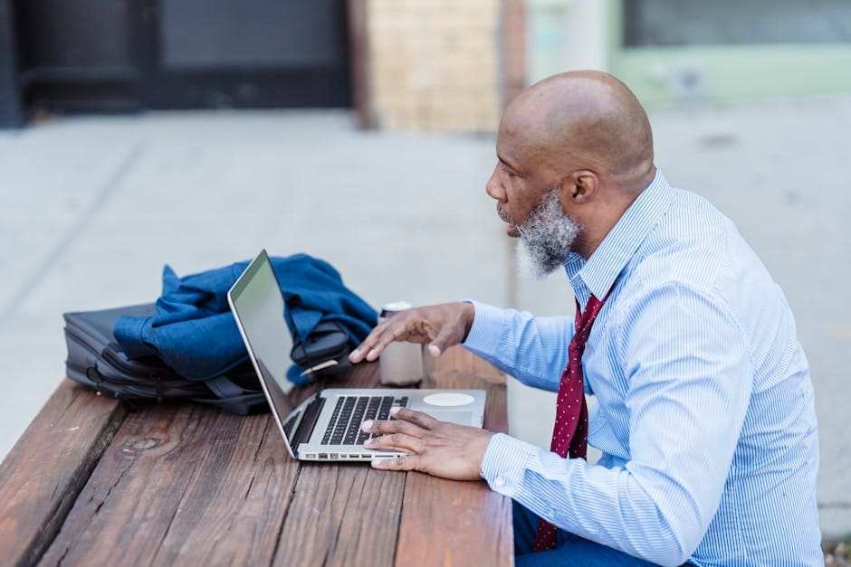 A professional man in a light blue shirt and red tie works on a laptop at an outdoor wooden table with a blue backpack beside him.