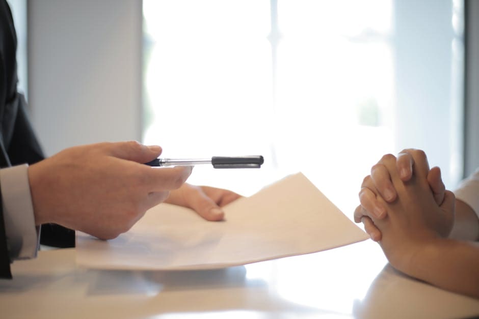 Two business professionals exchange or sign a contract document at a bright indoor workspace, symbolizing agreement or partnership.