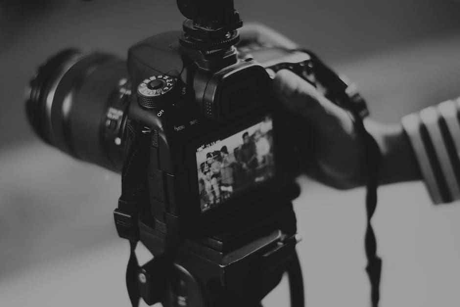 A close-up black and white photograph of a professional video camera being held, showcasing its equipment and technical details.