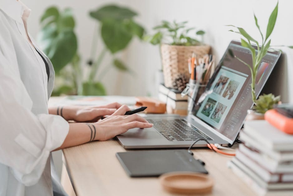 A person wearing a white shirt works at a modern desk with a laptop, surrounded by potted plants and office supplies in a bright, organized workspace.