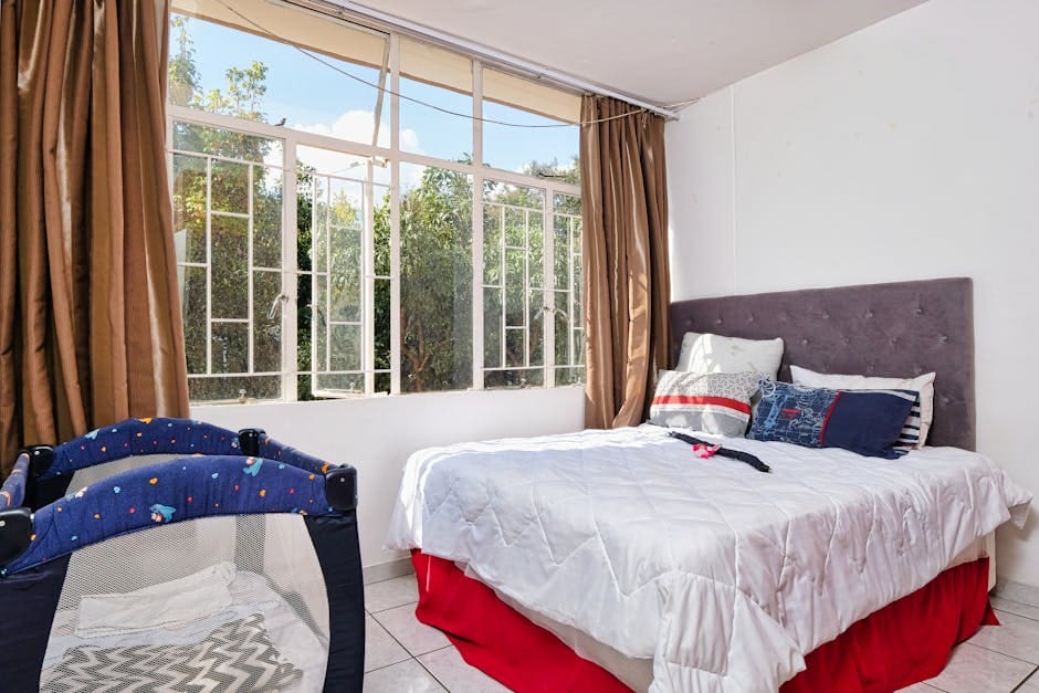 A bright, modern bedroom with a king-size bed and large windows overlooking a residential area, dressed in blue and red accents with neutral tones.