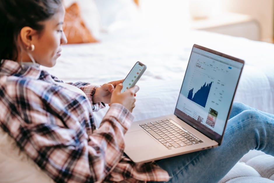 A person in a plaid shirt works remotely at a desk, using both a smartphone and laptop displaying data charts.
